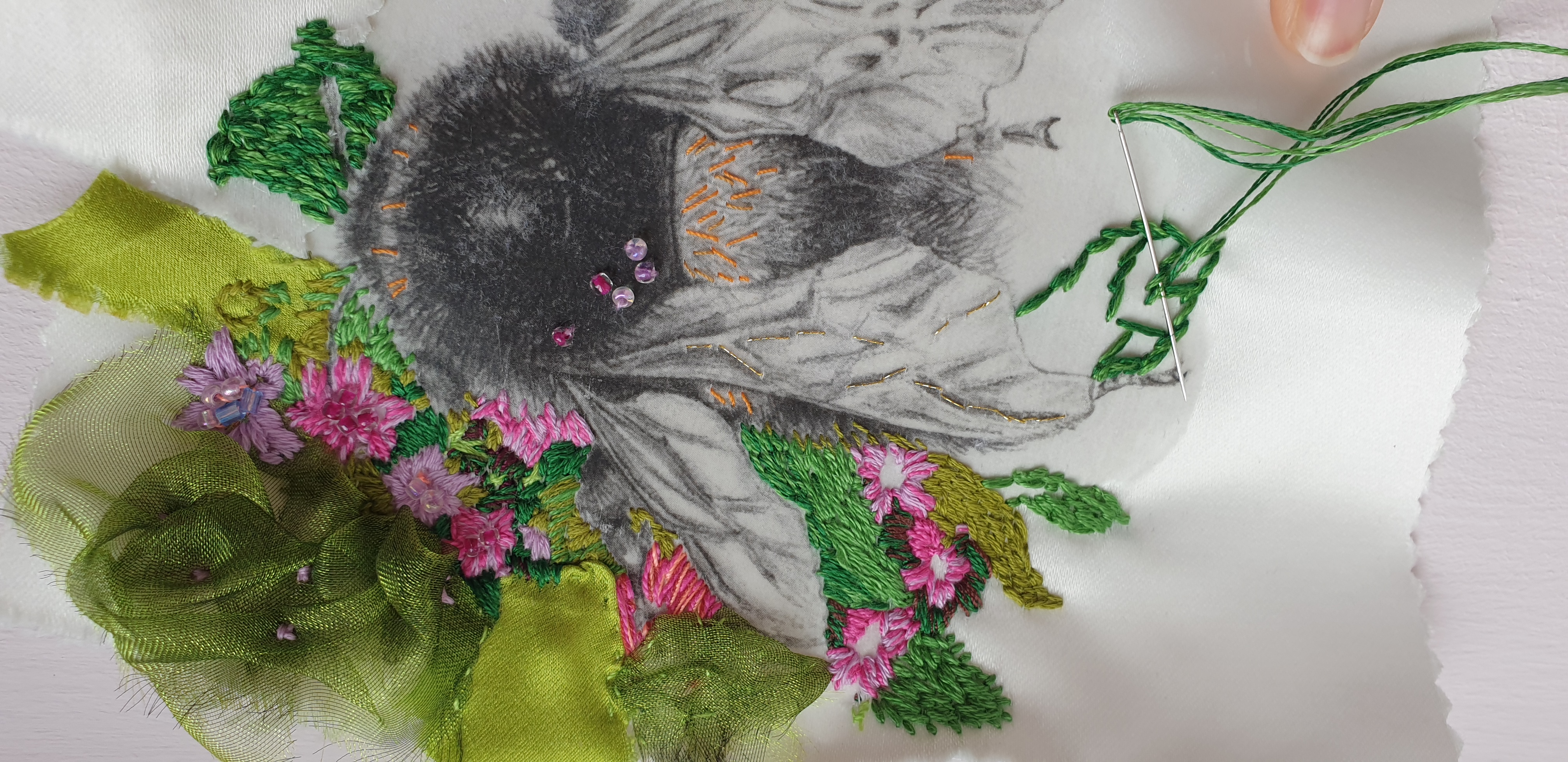 Photo of monochrome drawing of a bumblebee transferred onto white satin fabric surrounded by hand embroidered flowers and artist's hands embroidering leaves